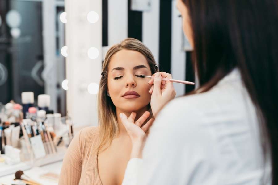 Maquilleuse appliquant des astuces maquillage sur les yeux d'une jeune femme blonde devant un miroir Hollywood illuminé.
