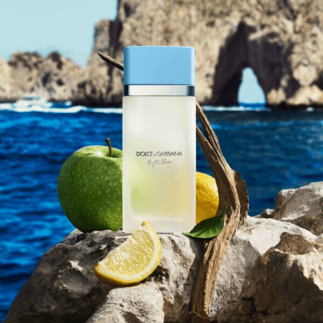 Flacon de parfum Light Blue de Dolce & Gabbana posé sur des rochers au bord de la mer Méditerranée, entouré d'une pomme verte et de citrons.