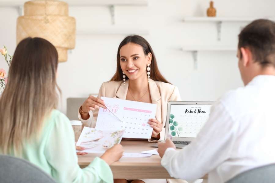 Un organisateur de mariage souriant présente un calendrier et des maquettes de papeterie à un couple assis à son bureau.