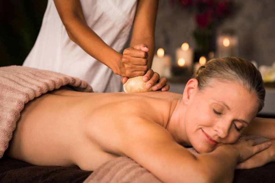 Gros plan montrant les mains expertes d'une praticienne massant le dos d'une femme avec un tampon de massage chaud lors d'un soin ayurvédique dans un spa.