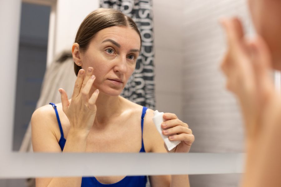 Une femme applique une crème de soin sur son visage en se regardant dans un miroir.